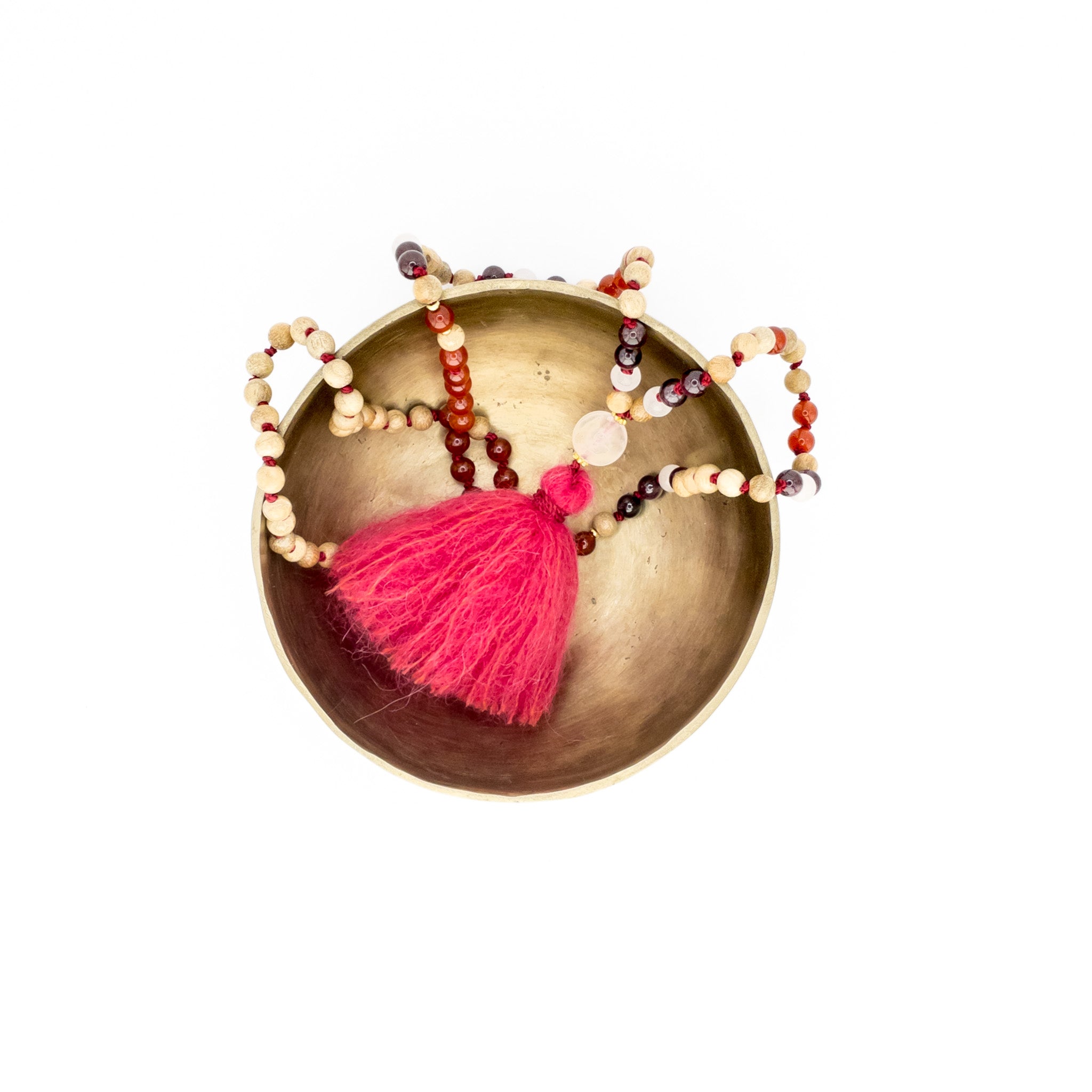 The Pink Petals Mala resting on a singing bowl, surrounded by other meditation and spiritual items. The mala is made with Rosewood beads, Carnelian, Garnet, and Pink Quartz beads, and is hand-knotted with a silk cord. The mala is adorned with a fluffy pink alpaca pompon and has a 12mm Ice Pink Quartz Guru Bead. shasha swiss mala jewellery
